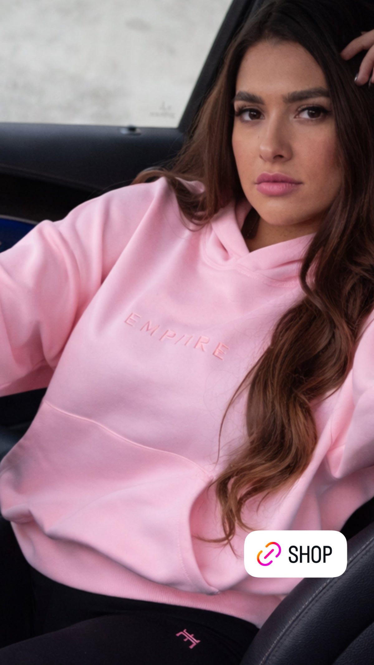 Woman wearing a pink hoodie with 'EMPIRE' branding, sitting inside a vehicle.