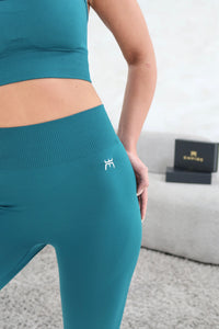 A close-up lifestyle shot of a model wearing a matching teal ribbed activewear set, featuring a sports bra and high-waisted leggings with a small white logo on the hip, set against a modern gray background.