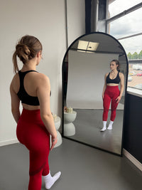 Woman in red leggings and black sports bra standing in front of a large mirror.