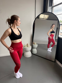 Woman in black sports bra and red leggings standing in front of a mirror in a room with a window.