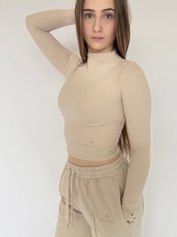 Woman wearing a beige long-sleeve top and pants with a visible brand logo on a white background