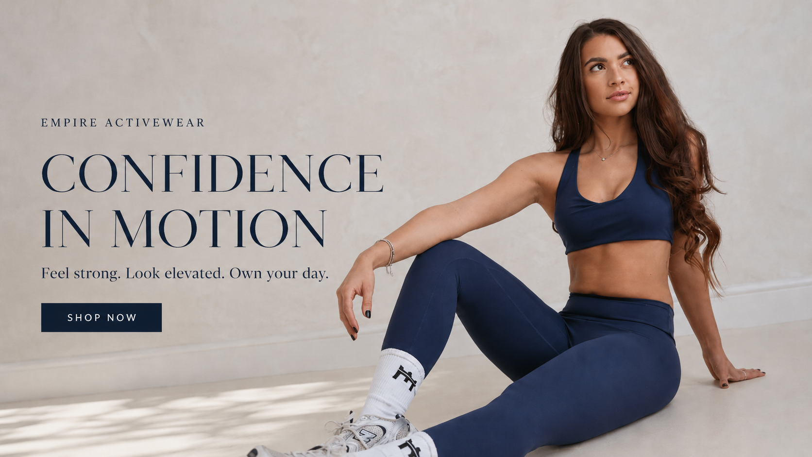 A woman wearing a navy blue sports bra and matching leggings sits on a light floor against a textured wall, with promotional text on the left reading 'EMPIRE ACTIVEWEAR CONFIDENCE IN MOTION' and a 'SHOP NOW' button.