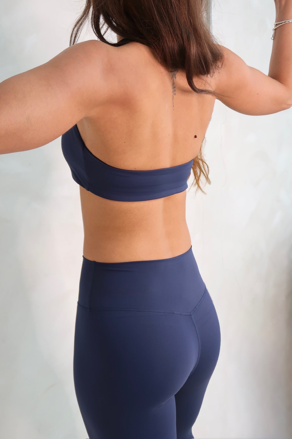 Rear view of a woman wearing a matching navy blue sports bra and high-waisted leggings set against a light textured wall.